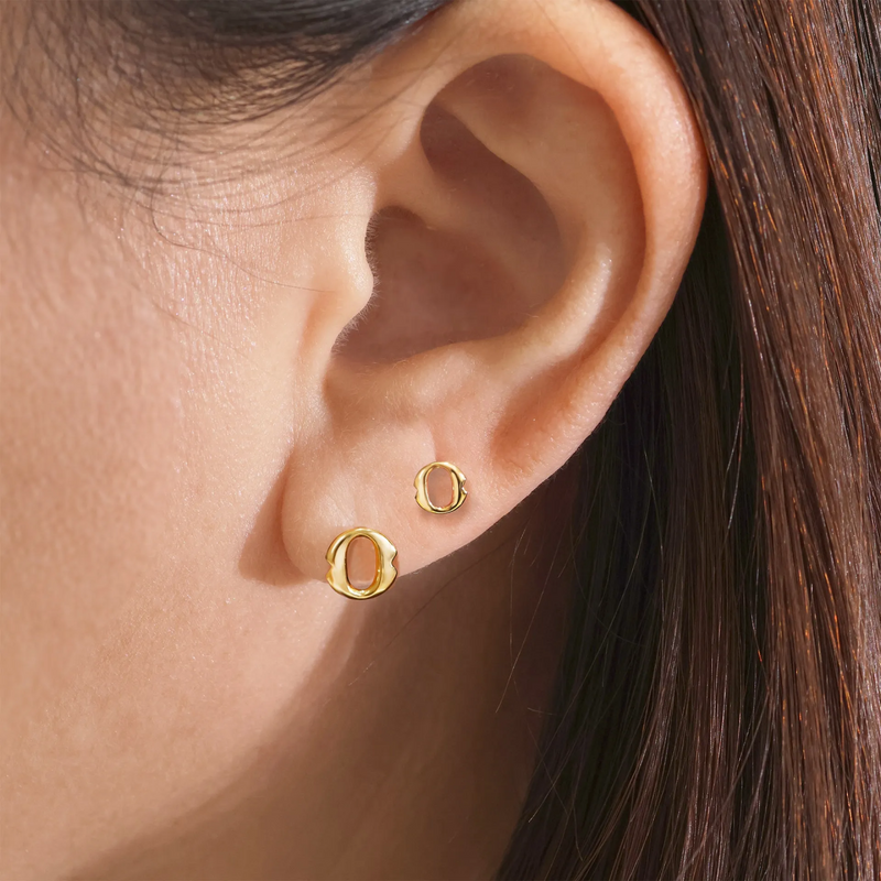 A pair of 18ct yellow gold small 'O' stud earrings from the Iconica Collection on the ear beside the smaller size to compare sizing