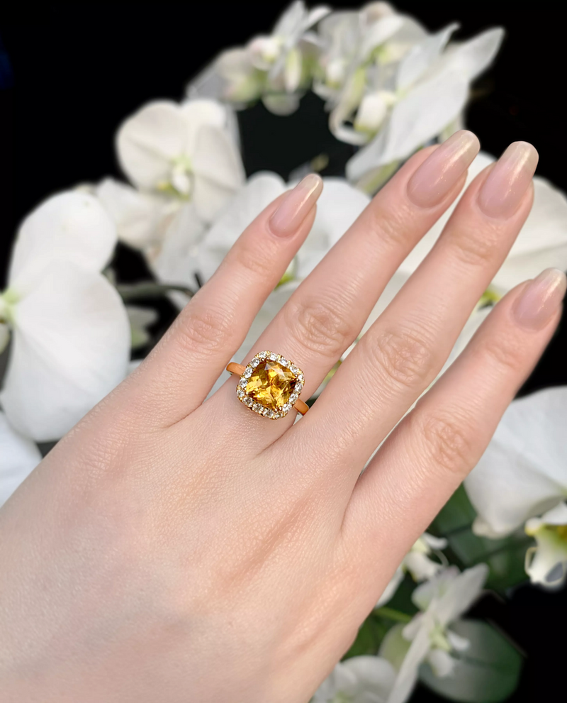 A cushion cut yellow tourmaline ring with round brilliant cut diamonds in a halo design, displayed on a person's hand with a blurred background of white flowers.